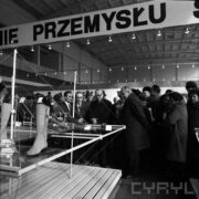 grupa mężczyzn, część z nich w płaszczach, część w garniturach, oglądają stoisko z damskimi butami w hali targowej