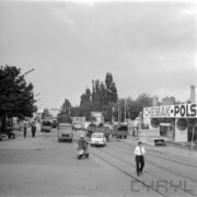 ulica na terenie targów, środkiem biegną tory, część samochodów osobowych i dostawczych zaparkowanych wzdłuż ulicy, część porusza się jej środkiem, na pierwszym planie mężczyzna na motorze