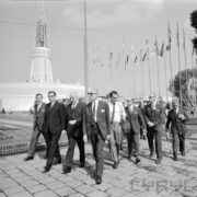 grupa spacerujących mężczyzn, z lewej budynek z iglicą, z prawej maszty z flagami