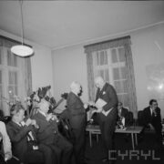 część mężczyzn siedzi przy stole, na stołach szklanki, kieliszki, butelki i talerze, część mężczyzn siedzi na krzesłach z lewej strony, w środku dwóch mężczyzn stoi