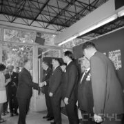 grupa ludzi w pawilonie wystawowym, jeden z mężczyzn wita się z pozostałymi