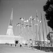 maszty z flagami państw, w głębi okrągły budynek z ażurową iglicą na dachu, spacerujący po placu ludzie