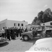 grupa mężczyzn w ogląda ekspozycję samochodów osobowych, w głębi otynkowany budynek