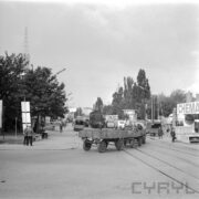 ulica na terenie targów, na pierwszym planie ciągnik z przyczepą, na której siedzi kilku mężczyzn, w głębi lokomotywa i samochody dostawcze, po obu stronach ulicy drzewa