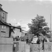 dwóch mężczyzn, jeden z nich słabo widoczny, w ubraniach roboczych ściąga z przyczepy samochodowej drewnianą skrzynię, którą podaje im mężczyzna, trzech mężczyzn, w tym dwóch w mundurach) stoi i przygląda się rozładunkowi, dookoła stoi kilka drewnianych skrzyń