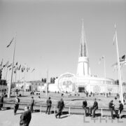 maszty z flagami państw i ażurową konstrukcją kuli ziemskiej pośrodku, w głębi okrągły budynek z ażurową iglicą na dachu, na placu spacerujący ludzie, część z nich siedzi na barierkach odgradzających teren targów