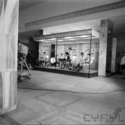 stoisko z kosmetykami, przy stołach na krzesłach siedzą kobiety, z lewej fragment kamery telewizyjnej