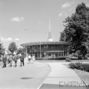 okrągły przeszklony budynek w otoczeniu drzew, za nim, w głębi ażurowa iglica, spacerujący ludzie