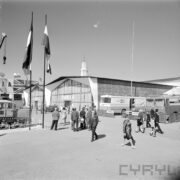 przeszklony budynek, zwiedzający ludzie, maszty z powiewającymi flagami