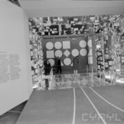 stoisko z przyklejonymi do szklanych ścian i sufitu pudełkami, zwiedzający ludzie, na wprost ściana z figurami geomatrycznymi