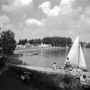 tory regatowe nad jeziorem, w głębi po trawie spacerują ludzie, na pierwszym planie drewniany pomost, na nim jedna kobieta i trzech mężczyzn w strojach kąpielowych, przy nich żaglówka