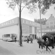 hala wystawowa, przed nią spacerujący ludzie, pod ściana hali zaparkowane dwa busy, z prawej stoisko z dużymi oponami