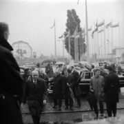 duża grupa ludzi w płaszczach, część pod parasolami, pomiędzy nimi samochody