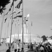 plac z masztami z flagami państw, w głębi okrągły budynek z ażurową iglicą na dachu, na placu spacerujący ludzie