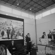 trzech mężczyzn przy maszynie w pawilonie wystawowym, na ścianie duże zdjęcie przywódcy koreańskiego w otoczeniu dzieci