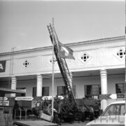 pośrodku dwóch mężczyzn podciąga flagę na maszcie, za nimi samochód z wyciąganą drabiną, jeden z mężczyzn stoi na samochodzie, za nimi fragment otynkowanego budynku z kolumnami