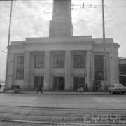 fragment białego budynku z ozdobnym portykiem z czterema kolumnami i trzema parami drzwi, nad drzwiami i po bokach duże okna, z przodu fragment ulicy z torowiskiem tramwajowym
