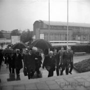 grupa mężczyzn w płaszczach, część z nich trzyma rozłożone parasole, wchodzi po schodach, za nimi murowany budynek z dużymi oknami