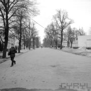 szeroka asfaltowa ulica na terenie targów, po bokach niej drzewa bez liści, z prawej i lewej pawilony targowe, z lewej idzie mężczyzna w kurtce i nakryciu głowy, w głębi spacerują ludzie