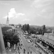 ulica z torami kolejowymi na terenie wystawy, dookoła budynki wystawowe, drzewa, spacerujący ludzie