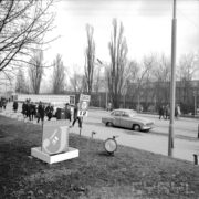aleja na terenie targów, po której przemieszczają się mężczyźni i kobiety, ubrani w płaszcze, większość w kapeluszach lub czapkach, przy ulicy stoją zaparkowane dwa samochody (jeden zasłaniają przechodnie), w głębi drzewa bez liści, na pierwszym planie na trawniku reklamy jubilera i pijalni czekolady