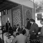 grupa kobiet i mężczyzn, część siedzi, a część stoi w pawilonie wystawowym, na ścianach powieszone dywany