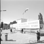 prostokątna tablica z napisem „witamy” w różnych językach na płocie wzdłuż ulicy, spacerujący ludzie