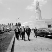 grupa mężczyzn w garniturach spacerujących po terenie targów, w głębi budynek z iglicą