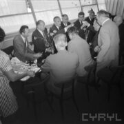 mężczyźni siedzą na krzesłach po obu stronach stolika, część z nich wznoszą toast kieliszkami do szampana, z lewej kobieta z tacą z kieliszkami