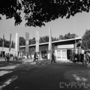 pawilon wystawowy, przed nim na masztach pionowe flagi i spacerujący ludzie