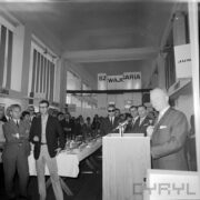 grupa ludzi w hali wystawowej, z prawej przy mównicy mężczyzna z mikrofonem, pośrodku hali zastawiony stół