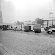 plac wyłożony betonowymi płytami chodnikowymi, w głębi samochody dostawcze, za nimi hale targowe