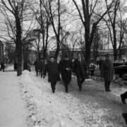 grupa mężczyzn w płaszczach, część z nich ma nakrycia głowy, idących chodnikiem, po bokach którego rosną drzewa, na poboczu leży odgarnięty śnieg