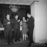 trzech mężczyzn i kobieta z kieliszkami w dłoniach, za nimi wisi żyrandol