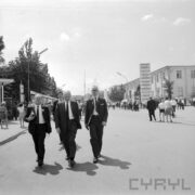 grupa kilku idących mężczyzn w garniturach, z prawej, w głębi murowany budynek z oknami