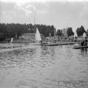 tory regatowe nad jeziorem, w głębi budynek ze skośnym dachem, spacerujący po trawie ludzie