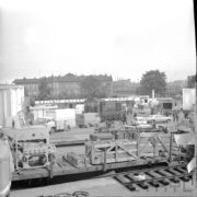 tereny targowe zastawione samochodami osobowymi, dostawczymi, w głębi na środku lokomotywa spalinowa, na pierwszym planie wagon ze skrzyniami