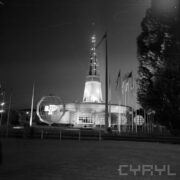 murowany budynek z iglicą, przed nim ażurowy model kuli ziemskiej z literką P, z prawej maszty z flagami państw, całość sfotografowana nocą