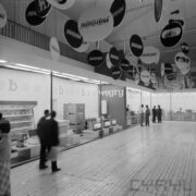 stoisko w hali wystawowej, zwiedzający ludzie, u sufitu wiszą okrągłe reklamy firm