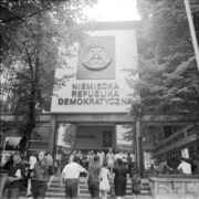 wejście do przeszklonego budynku, nad wejściem napis „Niemiecka Republika Demokratyczna”, po schodach do budynku wchodzą i wychodzą ludzie, po obu stronach wejścia drzewa