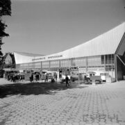 przeszklona hala wystawowa z wklęsłym dachem, przed halą stoiska z maszynami i samochodami, spacerujący ludzie