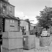 dwóch mężczyzn w ubraniach roboczych ściąga z przyczepy samochodowej drewnianą skrzynię, którą podaje im mężczyzna, jeden mężczyzna w furażerce przygląda się rozładunkowi, dookoła stoi kilka drewnianych skrzyń