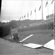 trawiasta skarpa, przy niej kilku mężczyzn montuje tablicę reklamową, nad skarpą maszty z flagami państw