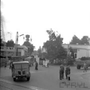 zaparkowane samochody dostawcze, grupki, stojących mężczyzn, po obu stronach ulicy budynki wystawowe, przed budynkiem z prawej drzewa