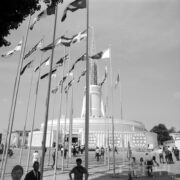 maszty z flagami państw, w głębi okrągły budynek z ażurową iglicą na dachu, na placu spacerujący ludzie