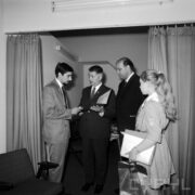 trzech mężczyzn, jeden z nich podaje drugiemu dyplom, z prawej dziewczynka w stroju harcerki