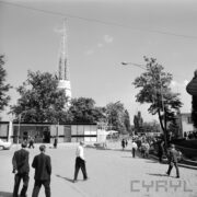 pawilony wystawowe, w głębi ażurowa iglica, spacerujący ludzie, wokół pawilonów drzewa