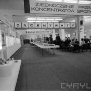 stoiska wystawowe w hali targowej, z prawej grupa kobiet i mężczyzn, siedzących na fotelach