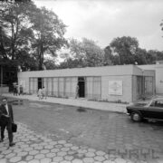 mały budynek murowany, z dużymi przysłoniętymi od wewnątrz oknami, spacerujący ludzie, z prawej fragment samochodu osobowego