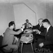 grupa mężczyzn siedzi przy stolikach, część z nich wznosi toast